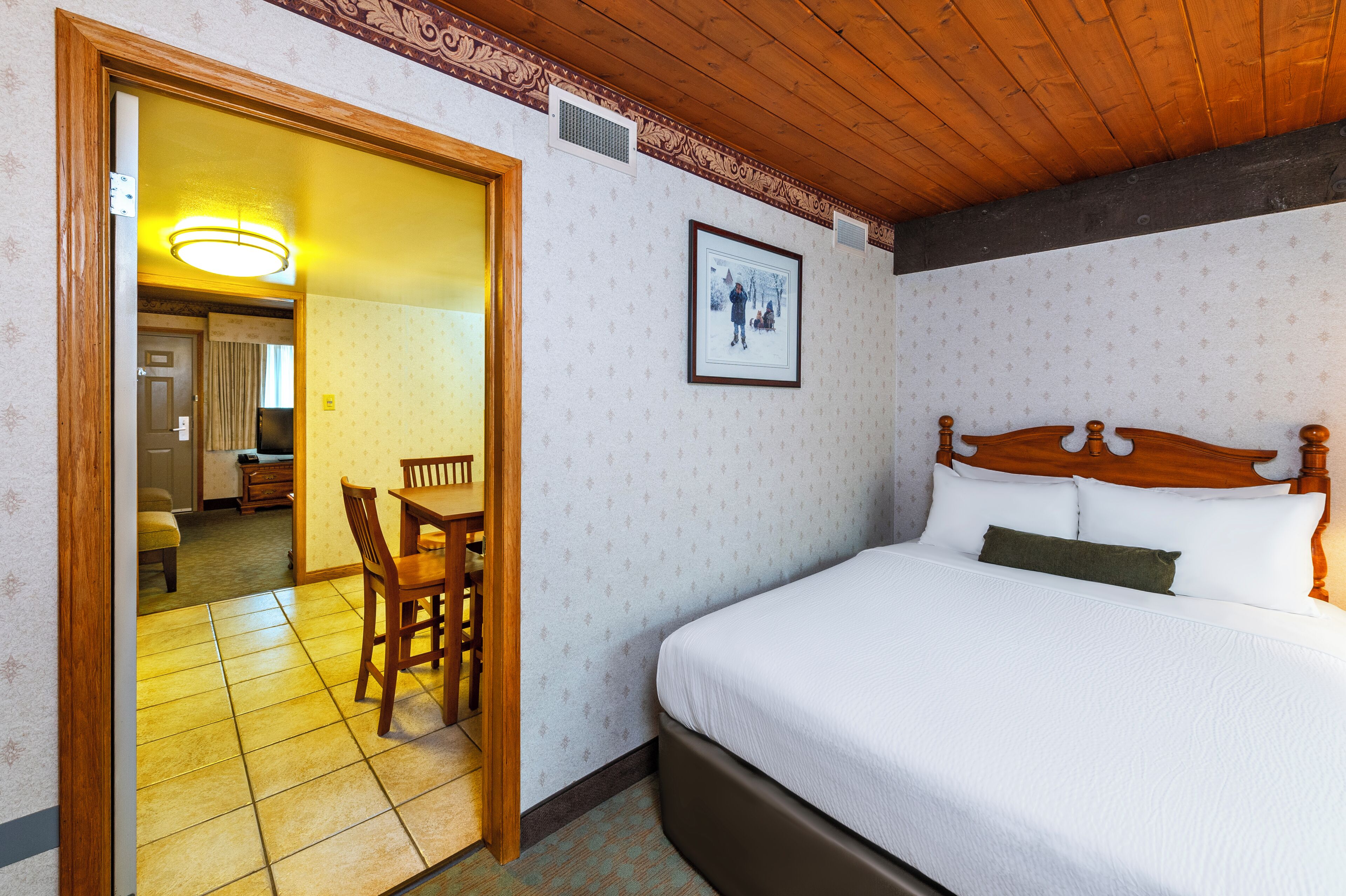 A hotel room with a bed, a dining table with chairs, a framed picture on the wall, and a wooden ceiling.