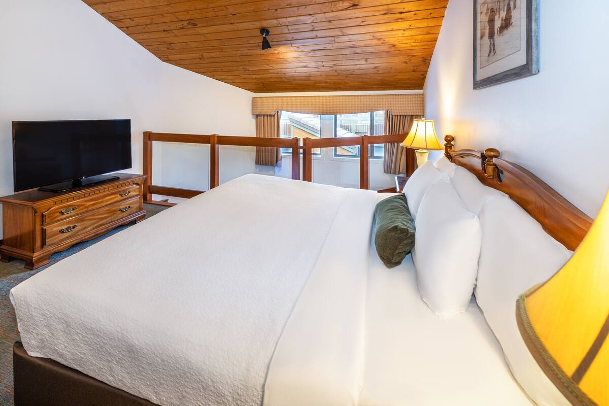 A bedroom with a wooden ceiling, a TV on a wooden dresser, a bed with white bedding, and a lamp on the nightstand.