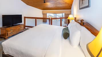 A bedroom with a wooden ceiling, a TV on a wooden dresser, a bed with white bedding, and a lamp on the nightstand.