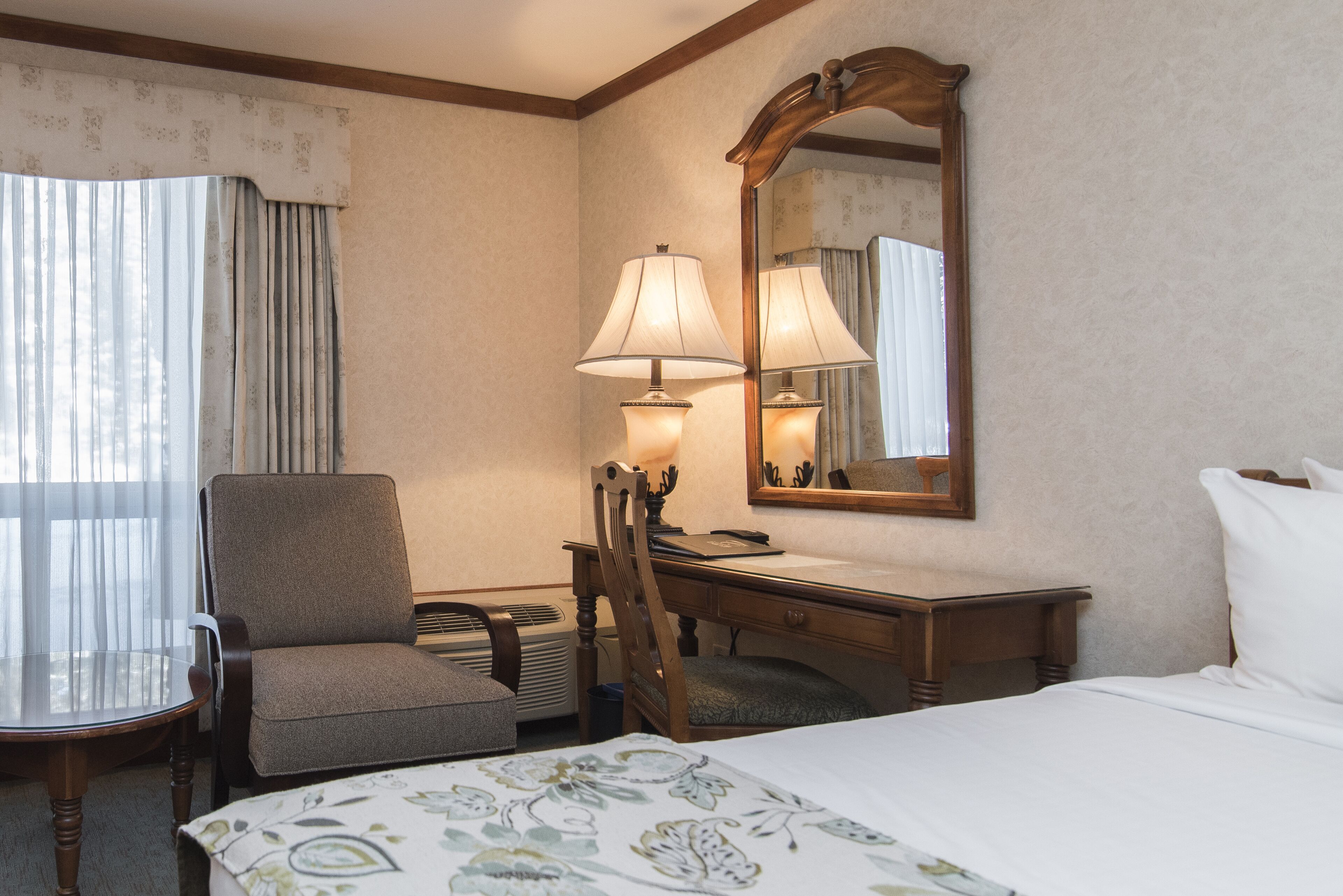 A hotel room with a bed, a desk, a chair, a lamp, and a large mirror.