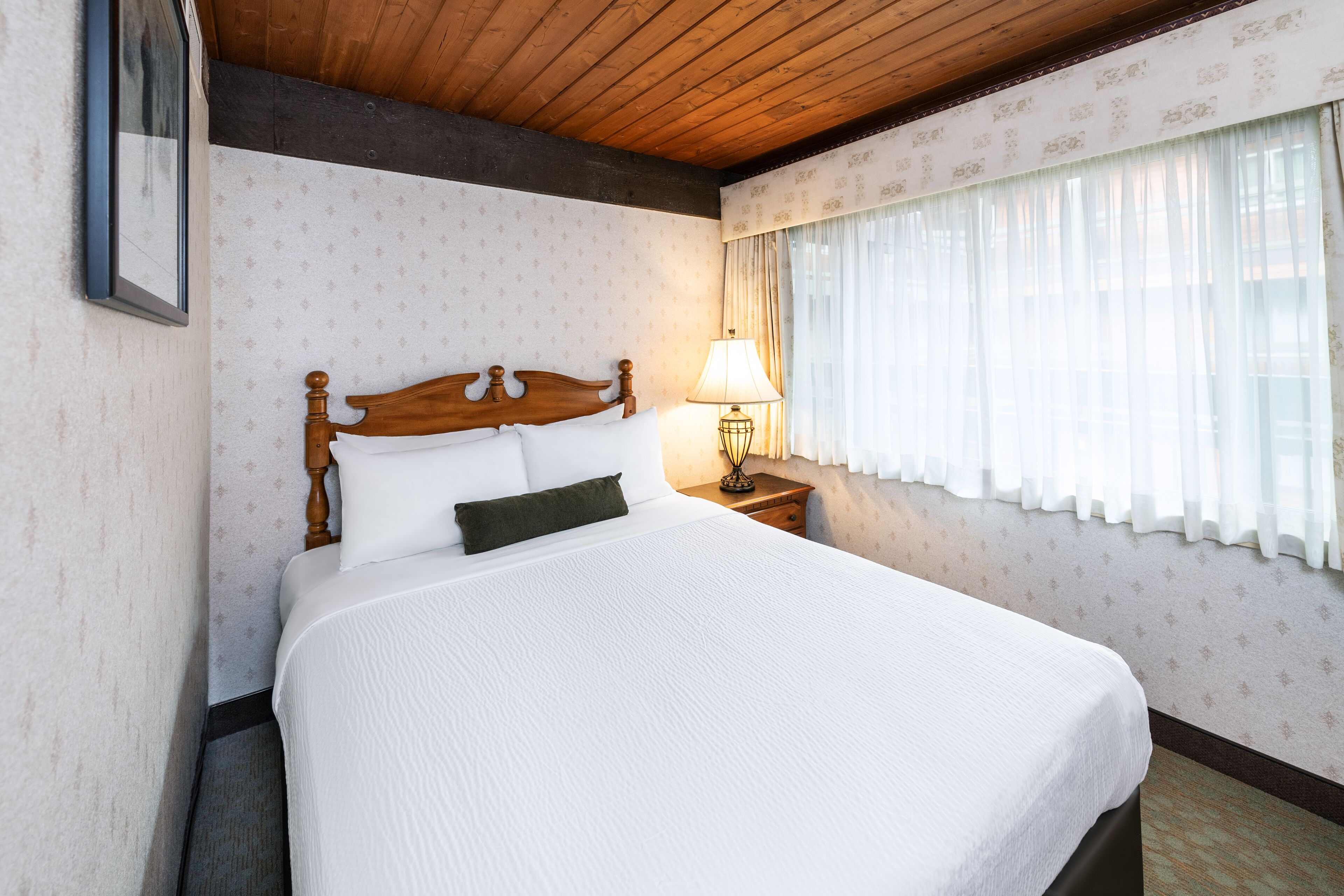Una cama bien hecha con cabecera de madera, lámpara de noche y ventana con cortinas traslúcidas.