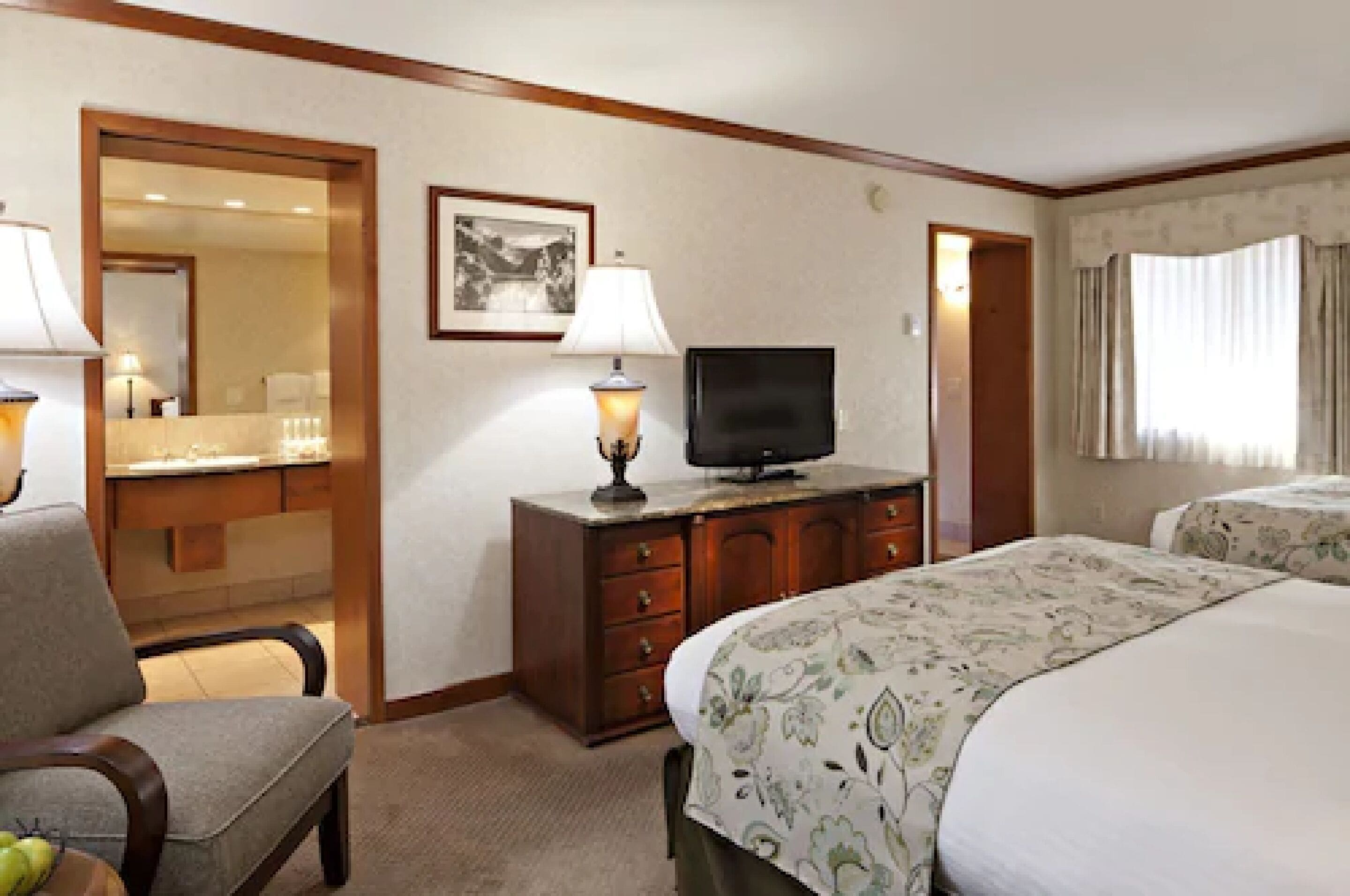 A hotel room with a bed, a television on a wooden dresser, a chair, and a desk with a lamp.