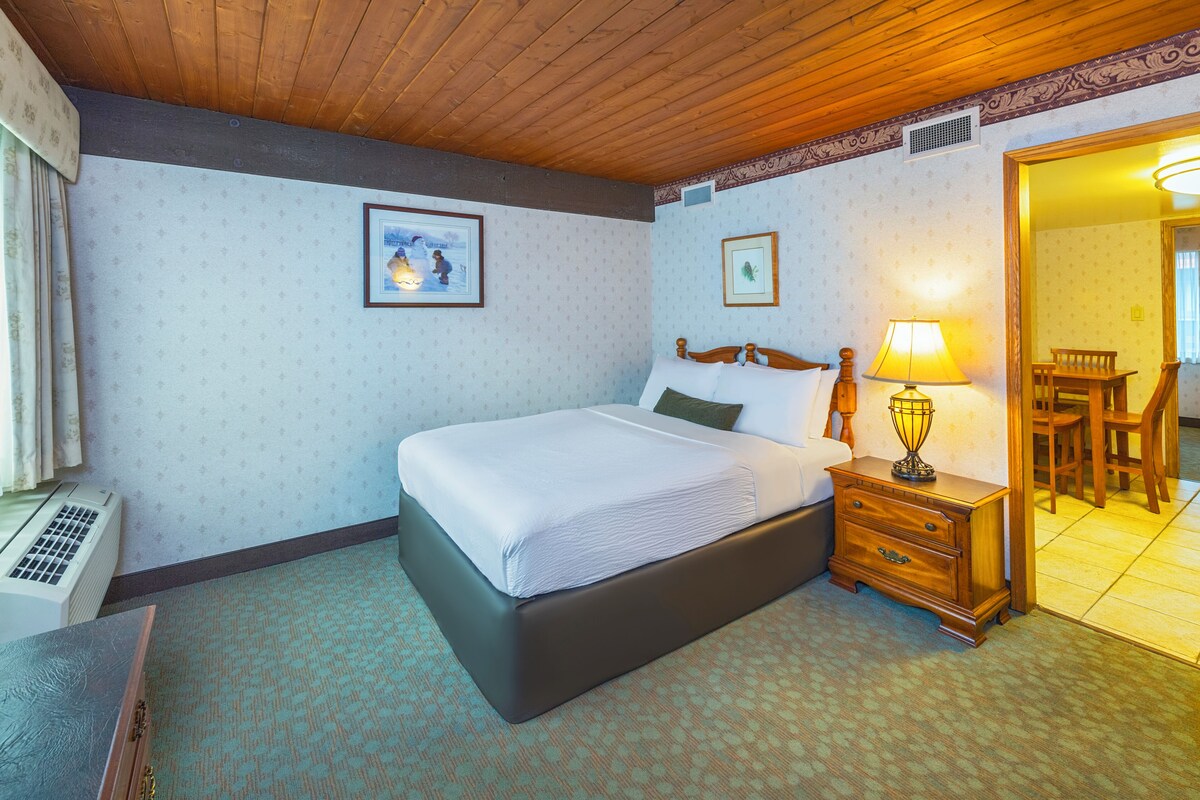 A hotel room with a large bed, a bedside table with a lamp, a framed picture on the wall, and a window with curtains.