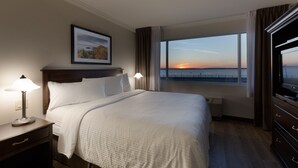 A hotel room with a large bed, a nightstand with a lamp, a TV, and a window with a sunset view. - Hotel Rimouski (Rimouski)