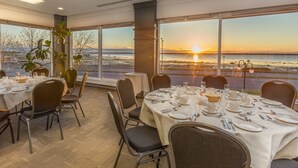 A dining area with tables set for a meal, large windows with a sunset view, and a plant in the corner. - Hotel Rimouski (Rimouski)