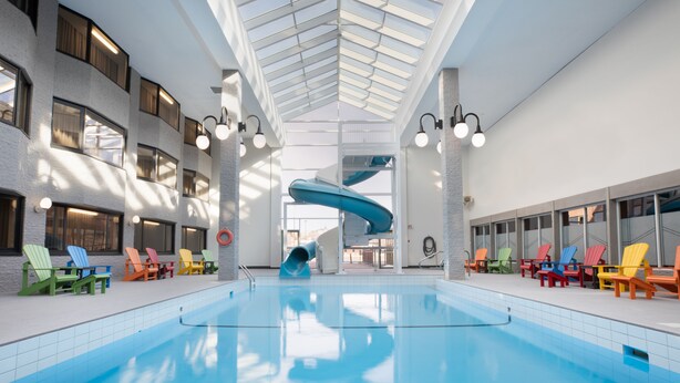 An indoor swimming pool with a blue slide, surrounded by colorful Adirondack chairs, and a high ceiling with skylights. - Hotel Rimouski (Rimouski)