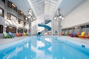 Una piscina interior con un tobogán azul, rodeada de sillas Adirondack de colores, y un techo alto con tragaluces.