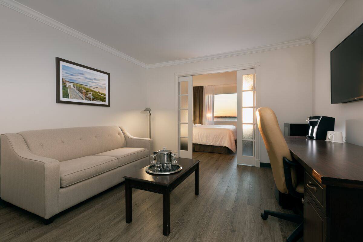 A hotel room with a sofa, a coffee table, a desk, a chair, a TV, and a bed with a view of the sunset.