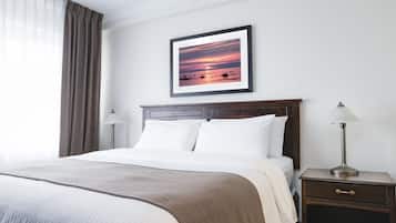 Una cama bien hecha con ropa de cama blanca y beige, un cuadro con una imagen de atardecer en la pared, una lámpara de noche y una mesita de noche de madera con un cajón.
