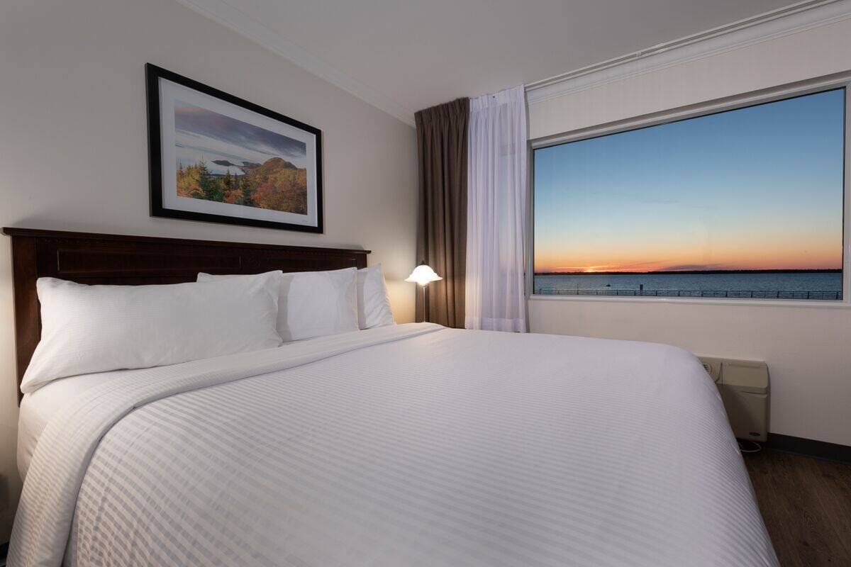 A hotel room with a large bed, a framed picture on the wall, a window with a view of the sunset, and a bedside table with a lamp.