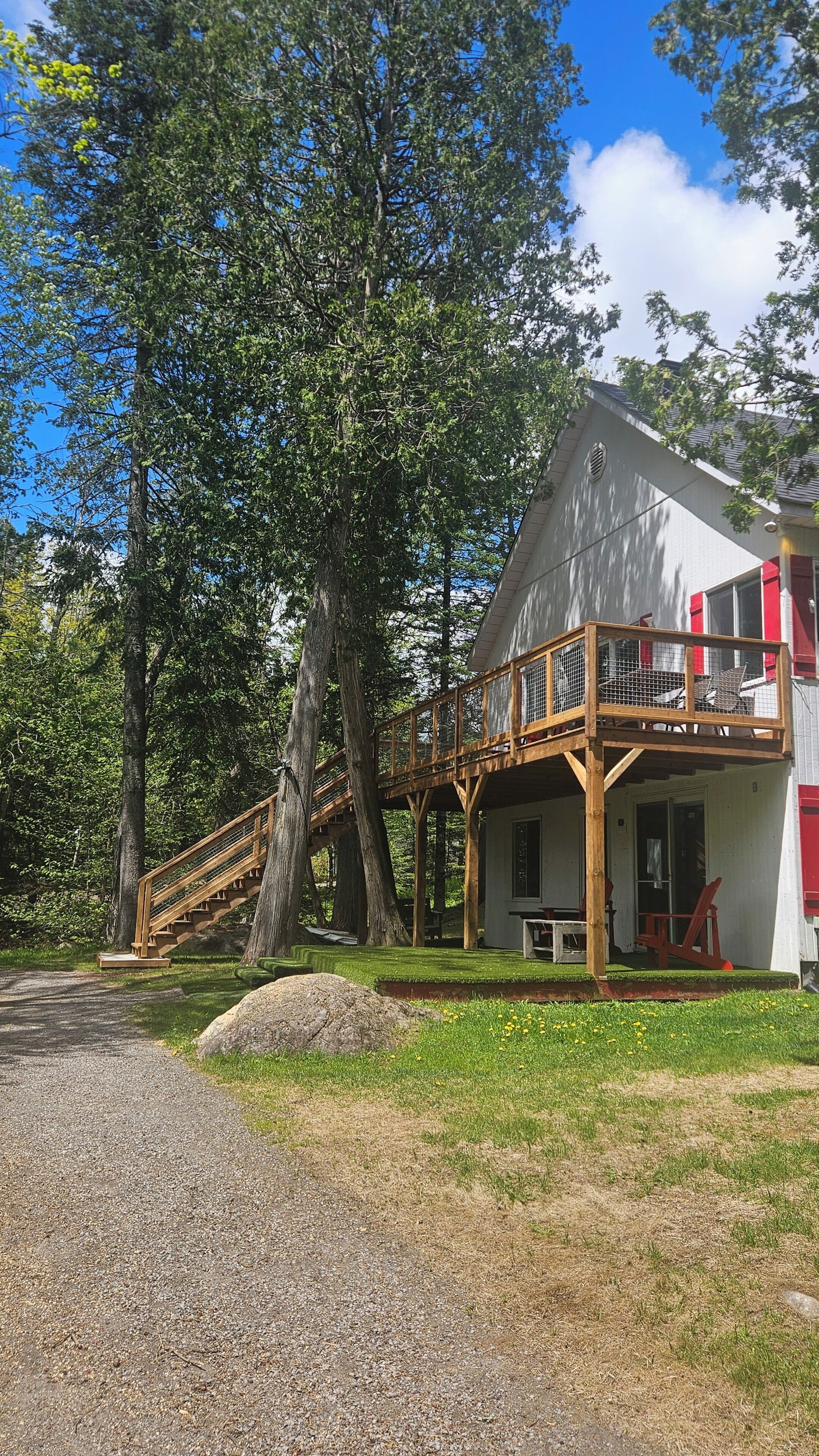 A two-story wooden house with a balcony, surrounded by trees and a gravel path.