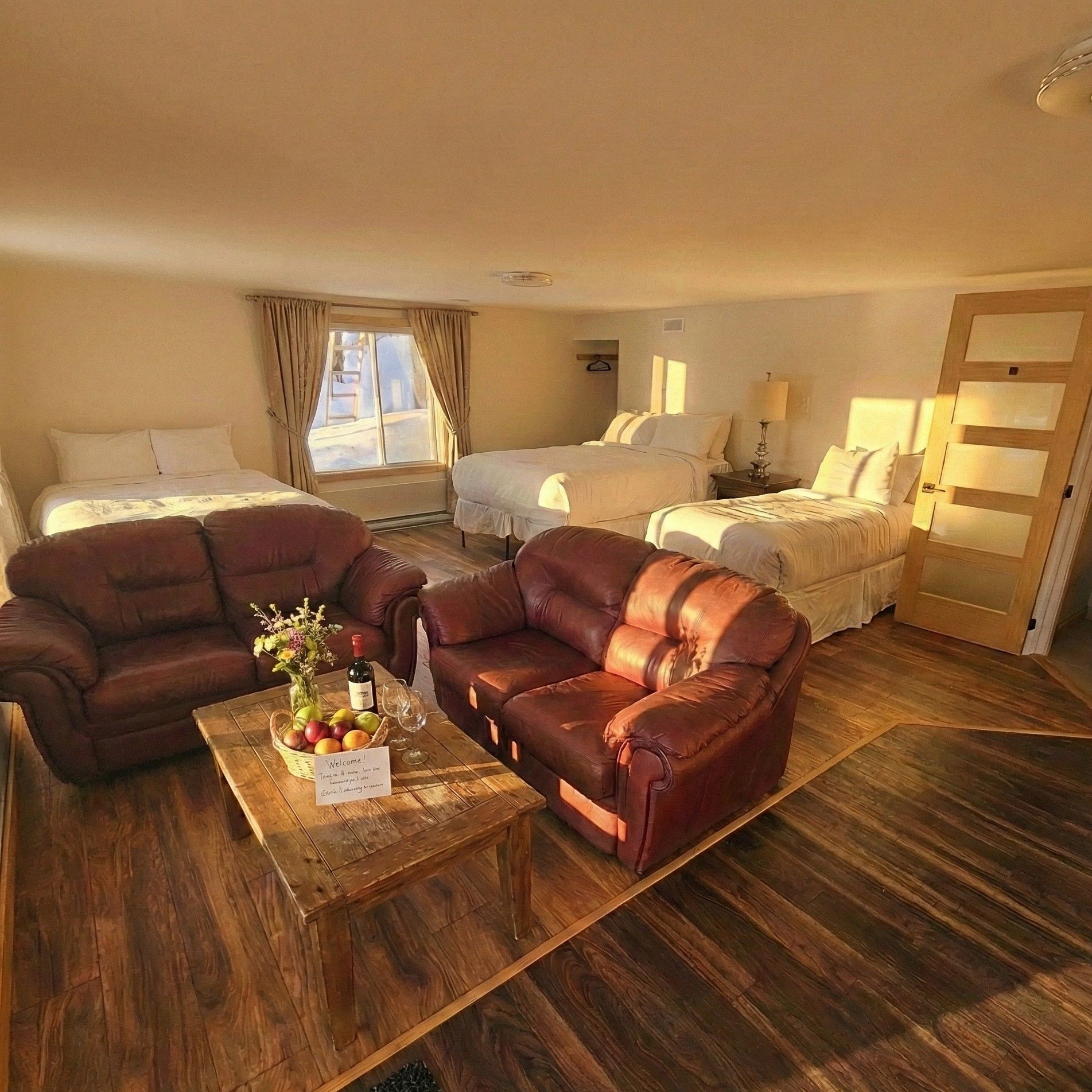 Ein Hotelzimmer mit zwei Betten, einem Sofa, einem Couchtisch mit Obst und einer Flasche sowie einem Holzboden.