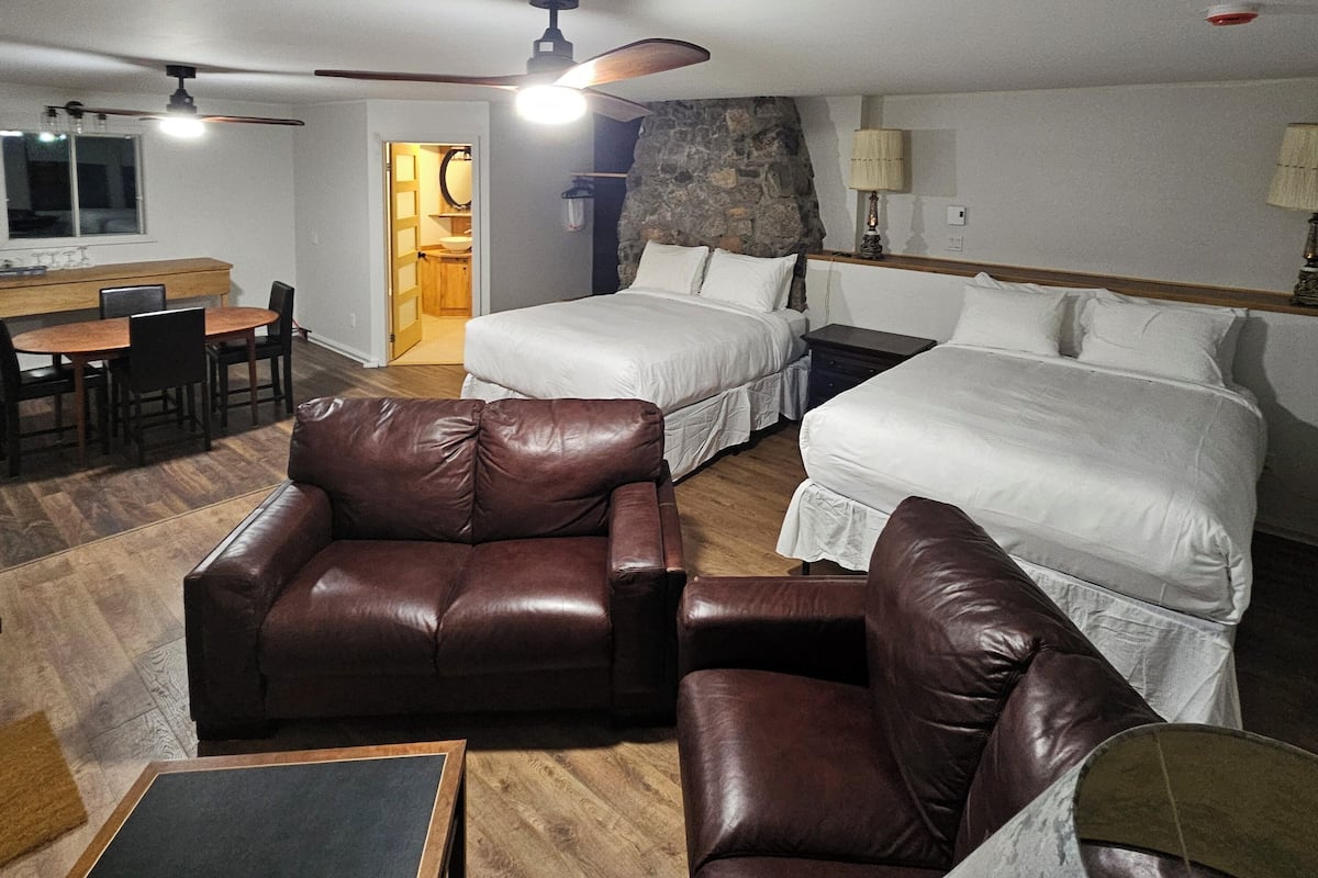 Quarto de hotel com duas camas, área de refeições, sofá de couro e parede com detalhes em pedra.