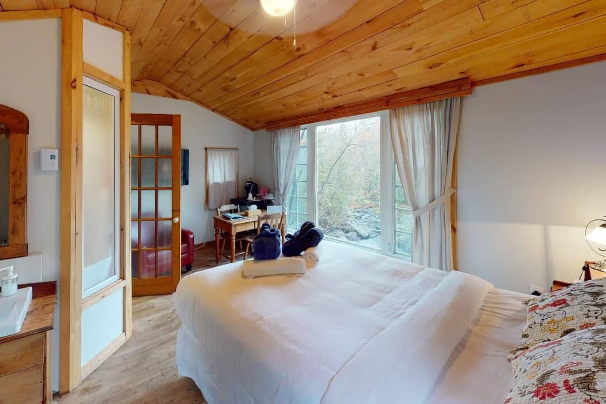 Quarto com uma cama grande, teto de madeira, janela com cortinas, mesa de cabeceira e espelho.