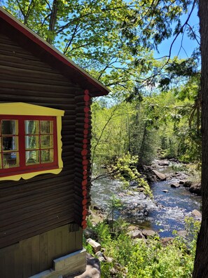 A log cabin with a red window and yellow trim, overlooking a rocky stream surrounded by dense forest. - Auberge & Spa Beaux-Rêves (Sainte-Adele)