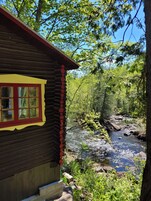 Una cabaña de troncos con ventana roja y detalles amarillos, con vistas a un arroyo pedregoso rodeado de un bosque denso.