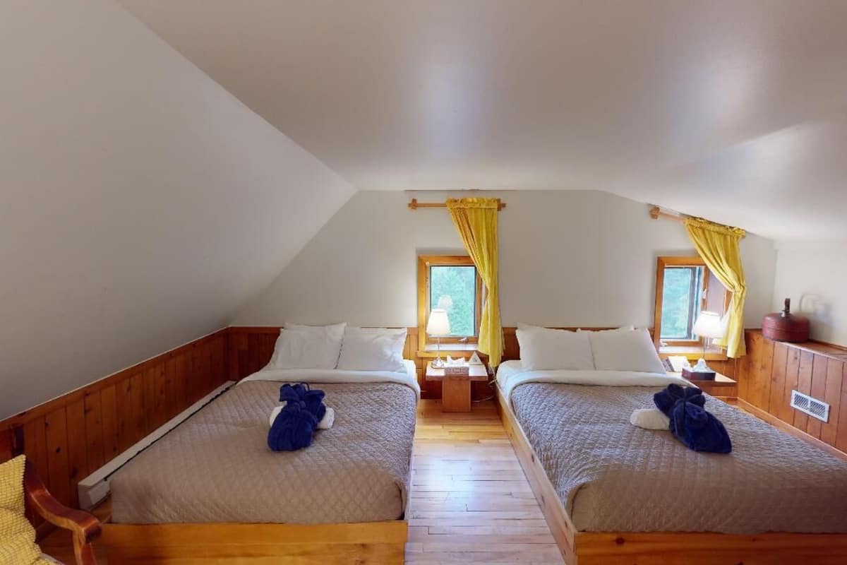 Quarto com duas camas, piso de madeira, janela com cortinas amarelas e uma pequena mesa entre as camas.