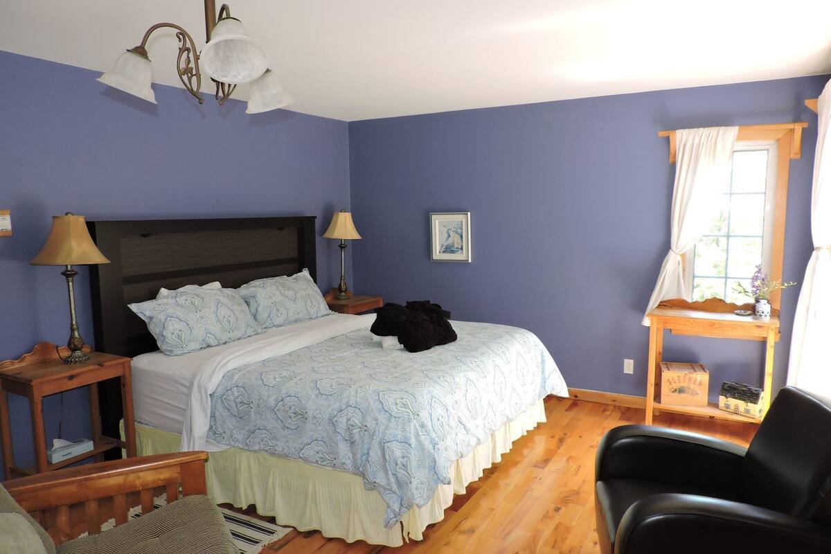Quarto com cama, duas mesas de cabeceira, uma cadeira, um banco de madeira, janela com cortinas e uma luminária na parede.