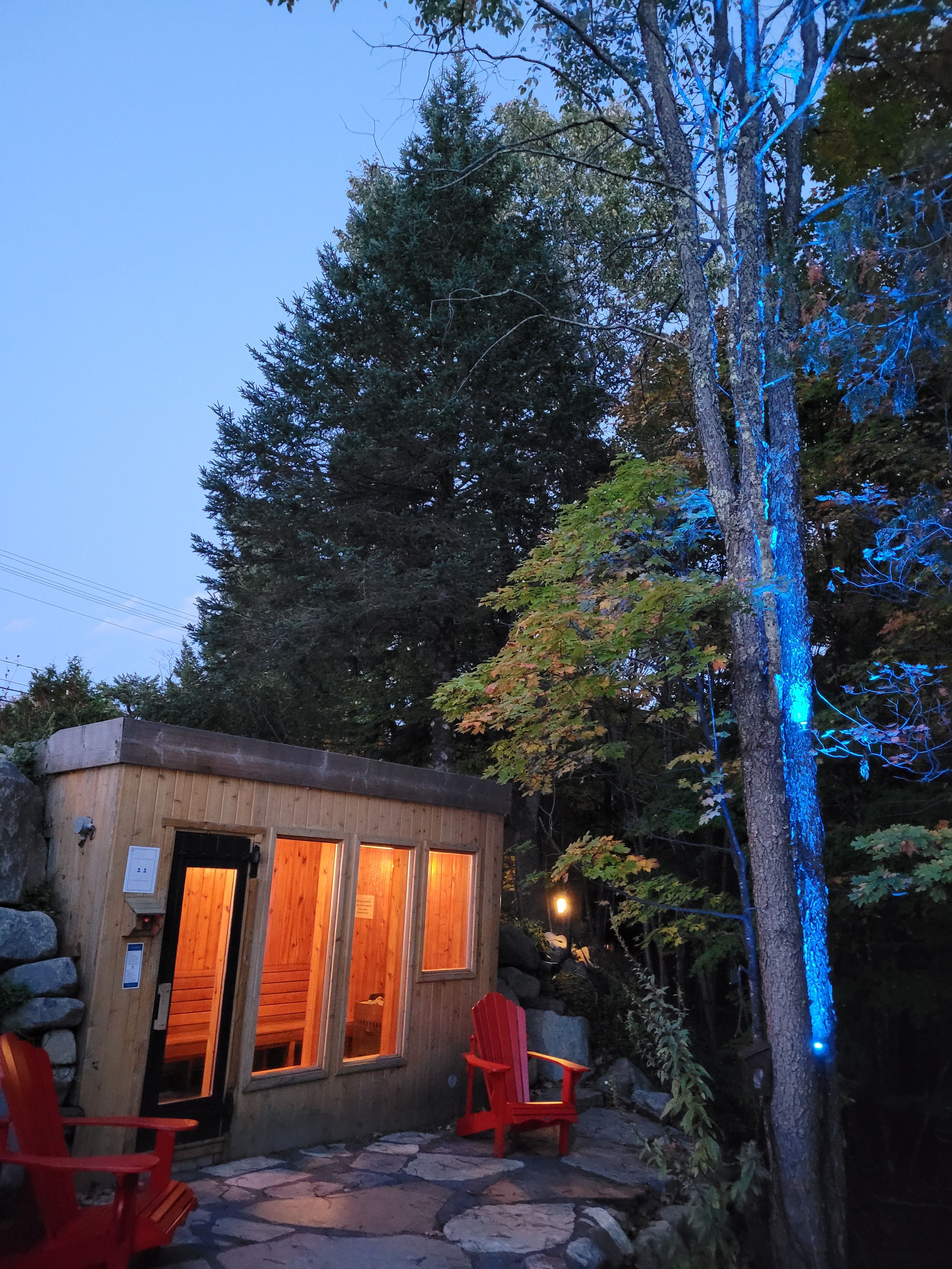 Een klein houten huisje met een steen terras, twee rode Adirondack stoelen en een boom met blauwe lampjes.