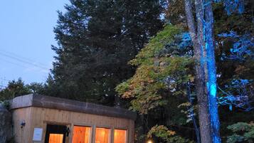 Een klein houten huisje met een steen terras, twee rode Adirondack stoelen en een boom met blauwe lampjes.