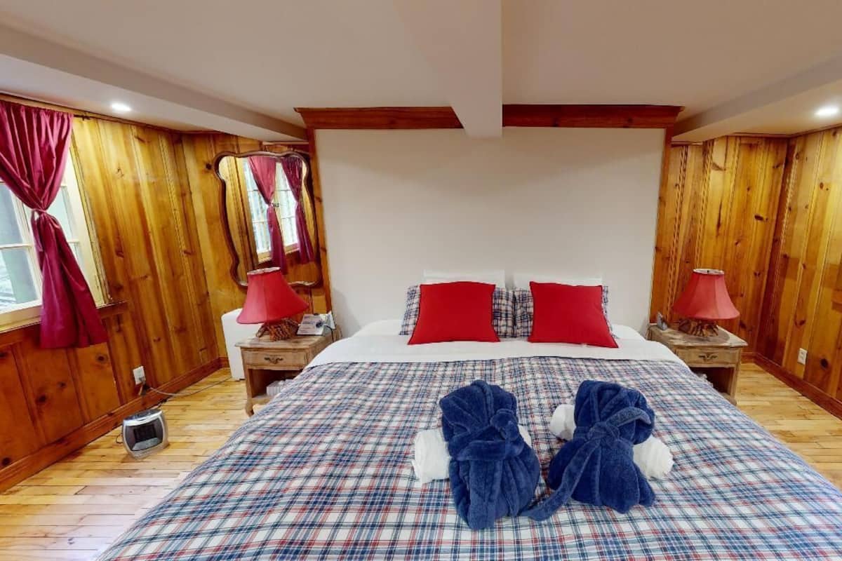 Quarto com piso de madeira, colcha xadrez, dois travesseiros vermelhos e dois roupões azuis na cama.
