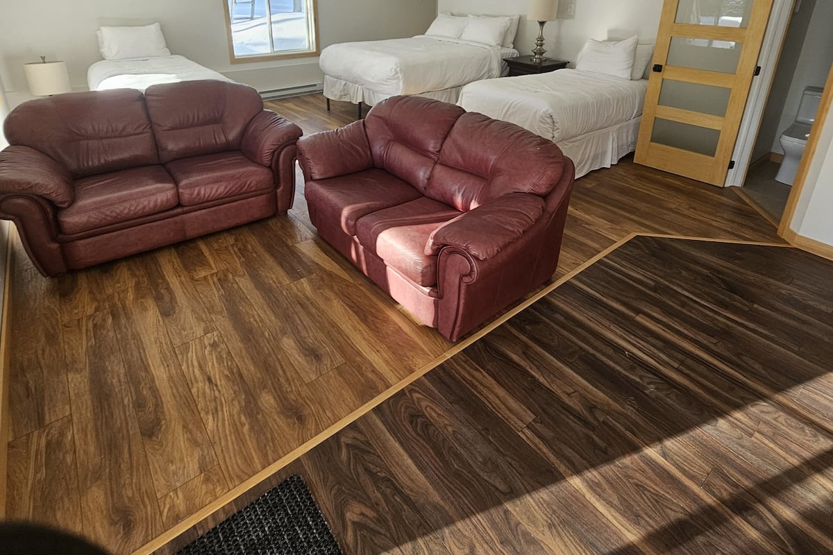 Quarto com duas camas, piso de madeira, sofá de couro vermelho, janela com vista e uma porta.