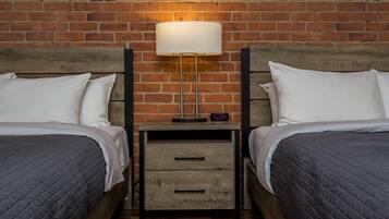 Habitación de hotel con dos camas, una mesita de noche con lámpara y una pared de ladrillo.