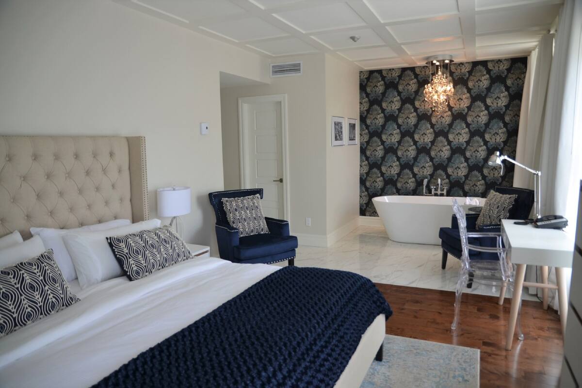 A bedroom with a large bed, a chair, a desk, and a bathroom with a bathtub and shower.