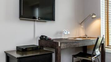 A hotel room with a flat-screen TV mounted on the wall, a desk with a lamp, a chair, a small cabinet, and a window with blinds.