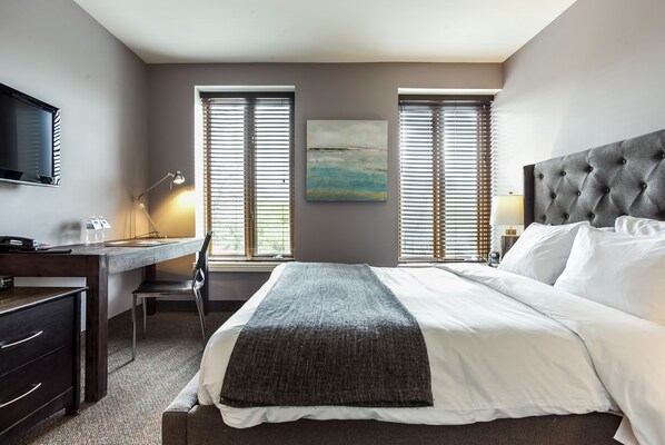 A hotel room with a bed, a desk, a chair, a television, and two windows with blinds.