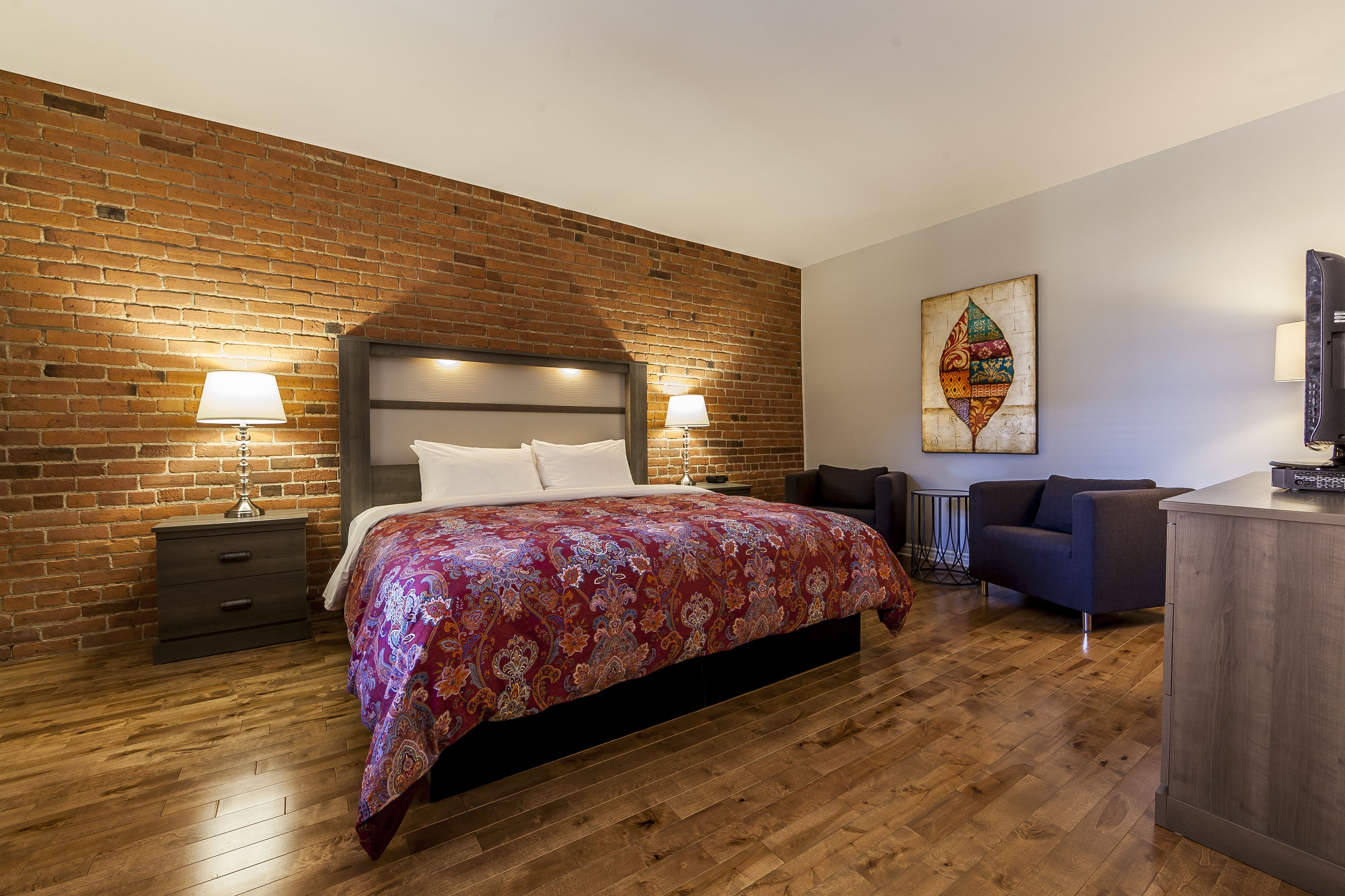 A bedroom with a brick wall, a large bed with a patterned bedspread, two bedside lamps, a desk with a chair, and a decorative wall piece.