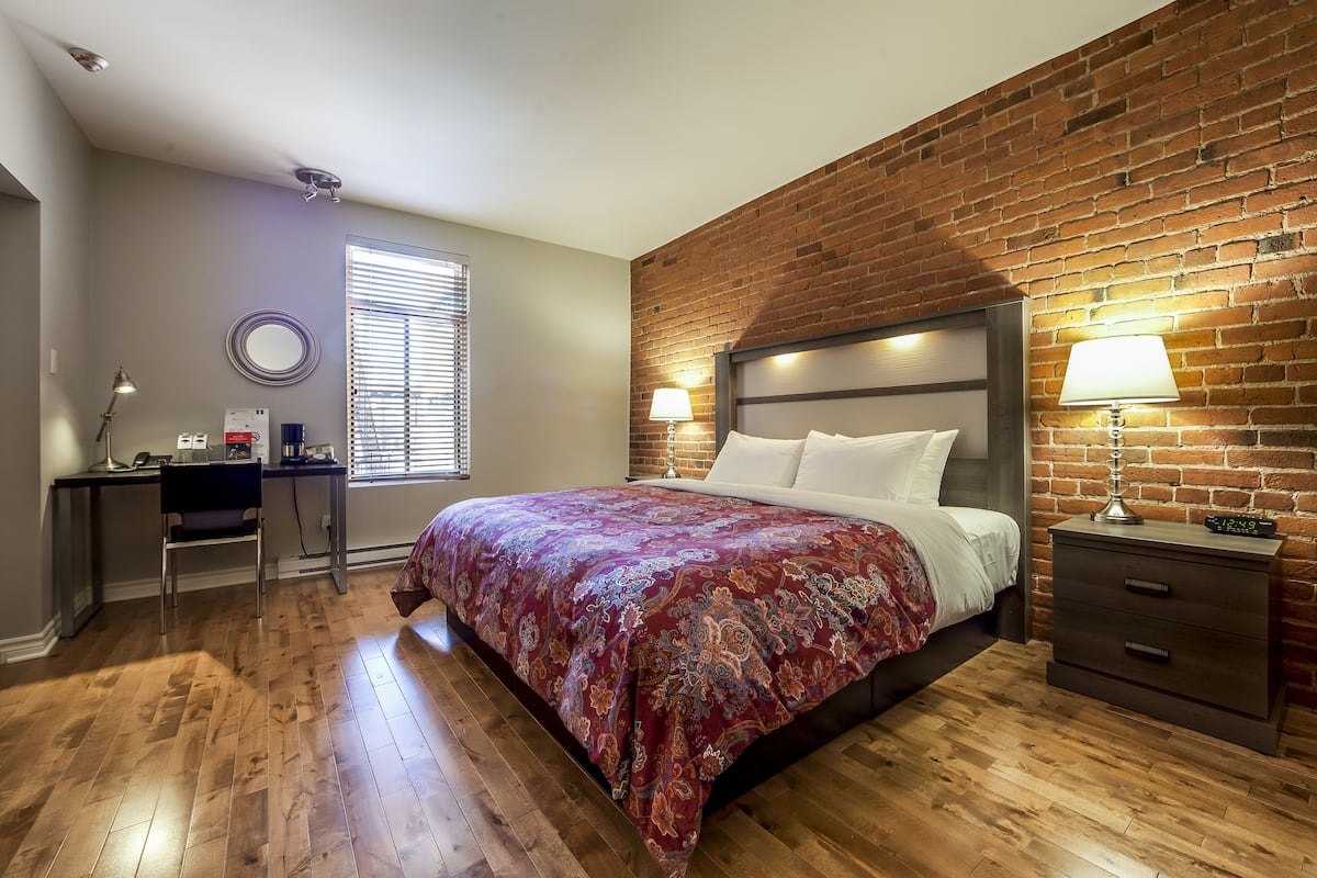 A bedroom with a brick wall, a bed with white linens, a desk with a chair, and a nightstand with a lamp.