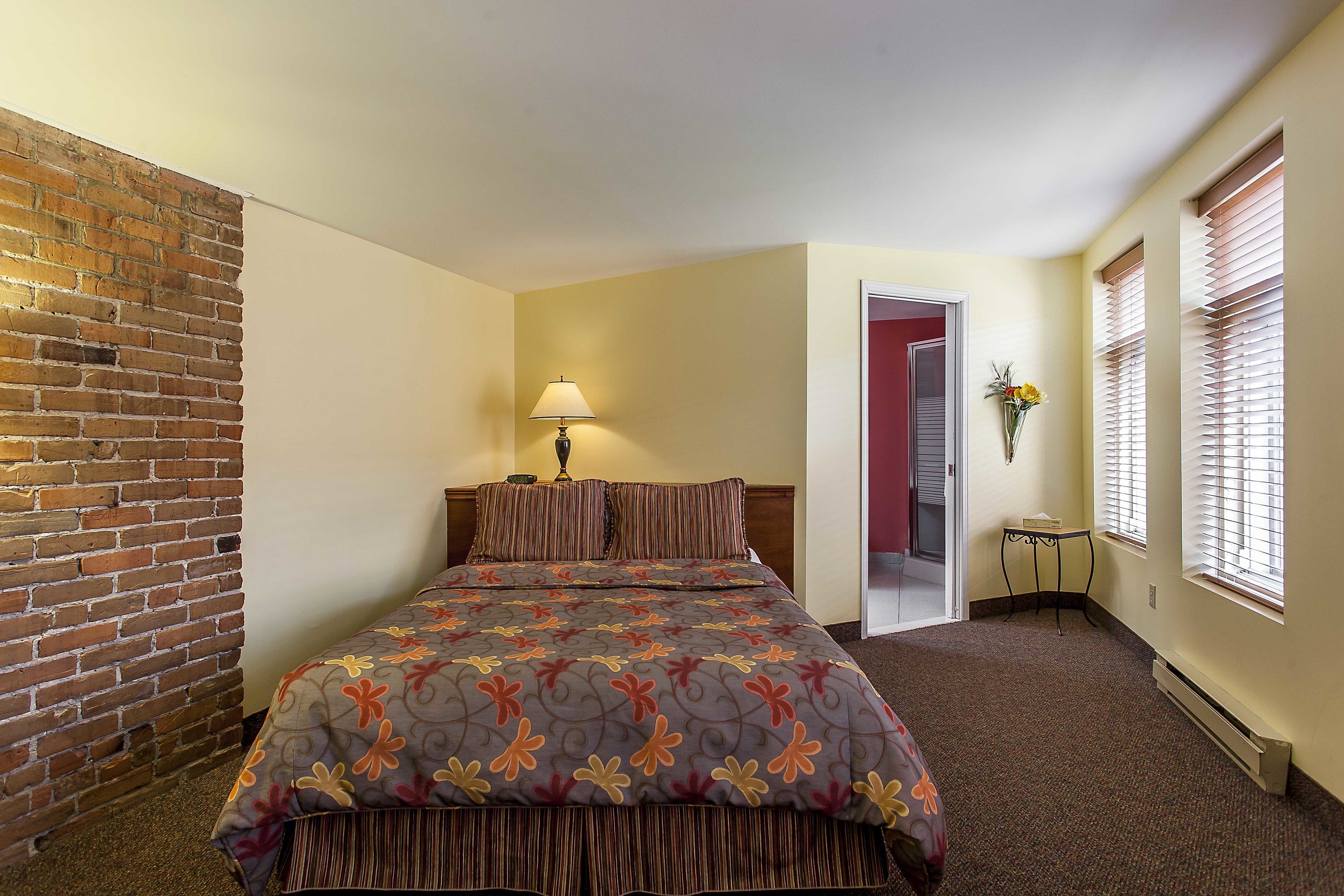 A hotel room with a brick wall, a bed with a floral bedspread, a bedside table with a lamp, and a window with blinds.