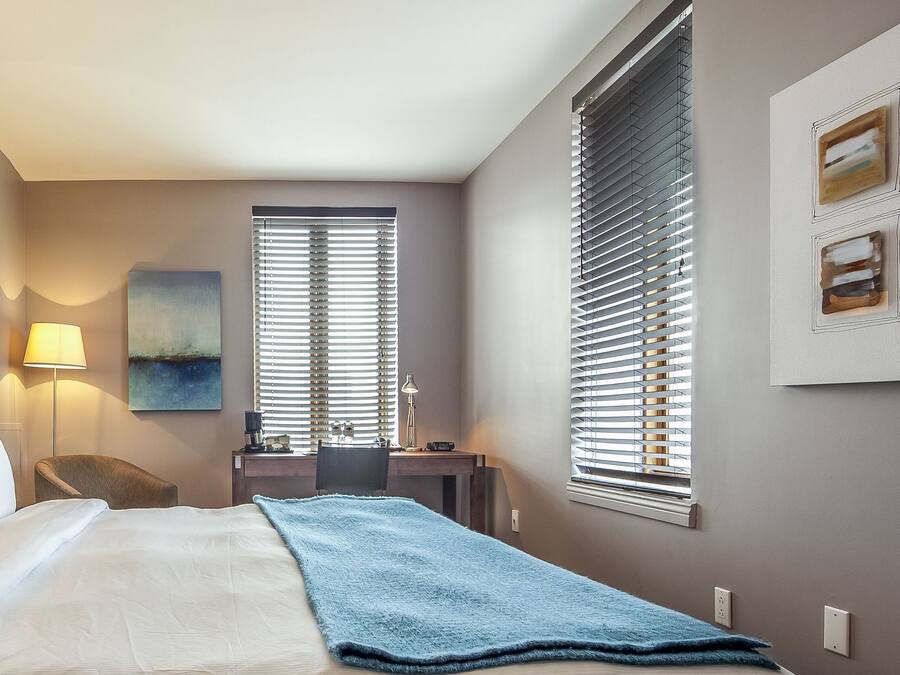 Une chambre d’hôtel avec un lit, deux lampes de chevet, un bureau avec une chaise, une fenêtre avec des stores et un tableau au mur.