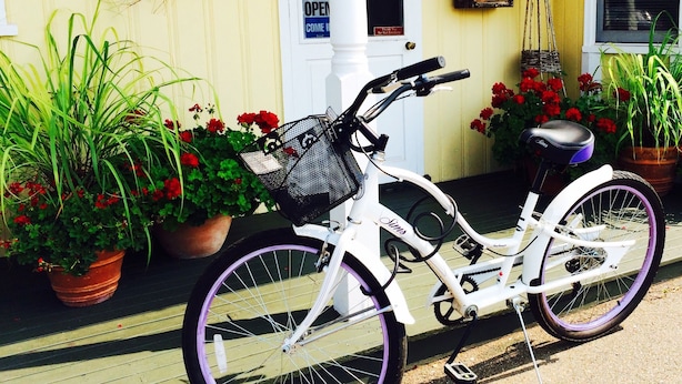 A white bicycle with a basket and a seat, parked in front of a yellow building with a wreath on the door, potted plants, and a sign that says "OPEN".