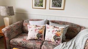 A living room with a floral-patterned sofa, two framed pictures on the wall, a lamp, and a magazine on a coffee table.