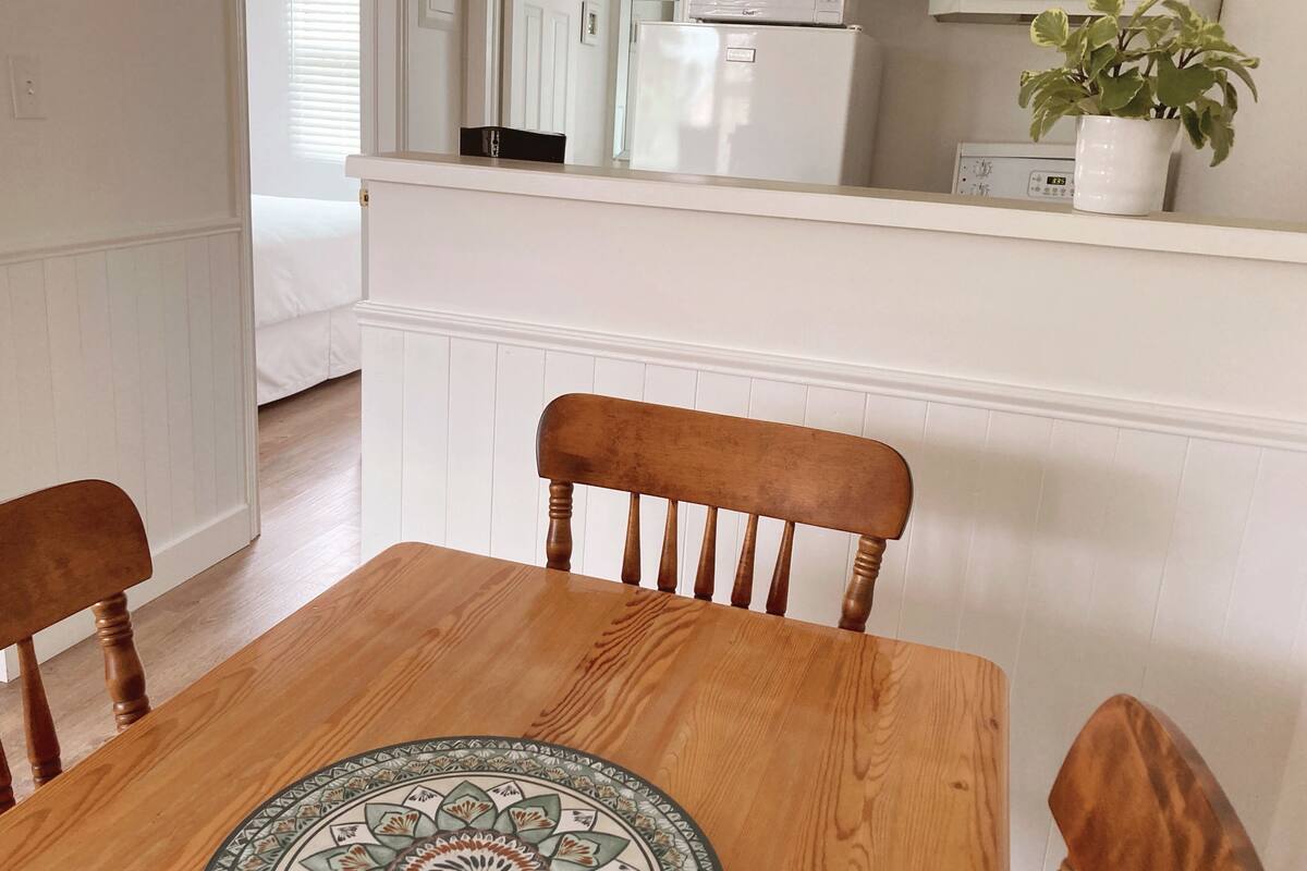 A dining area with a wooden table and chairs, a decorative rug, and a white kitchen in the background.