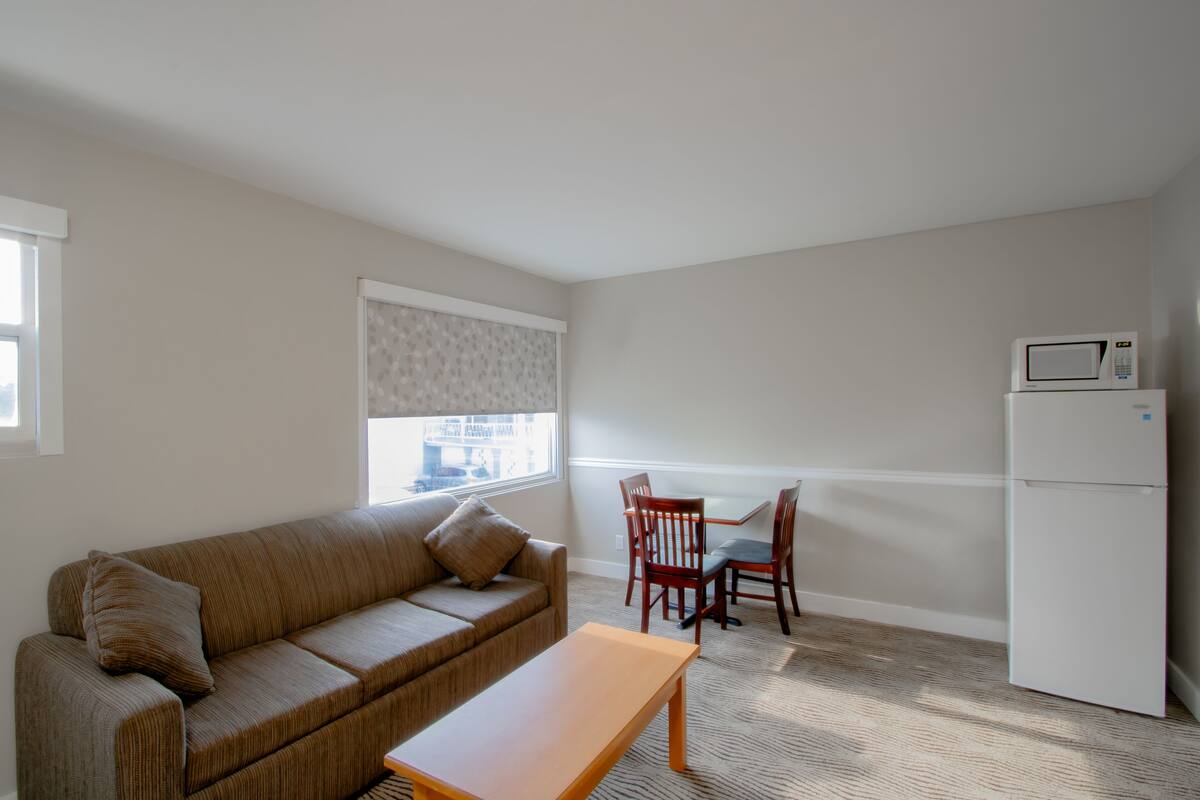 A compact living space with a sofa, a small dining table, and chairs. A refrigerator is visible in the corner.