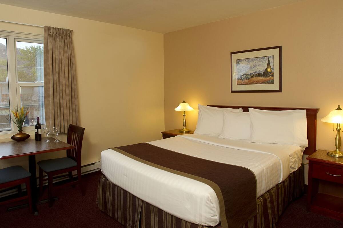 A hotel room with a bed, two bedside lamps, a desk with a chair, a vase with flowers, and a painting on the wall.