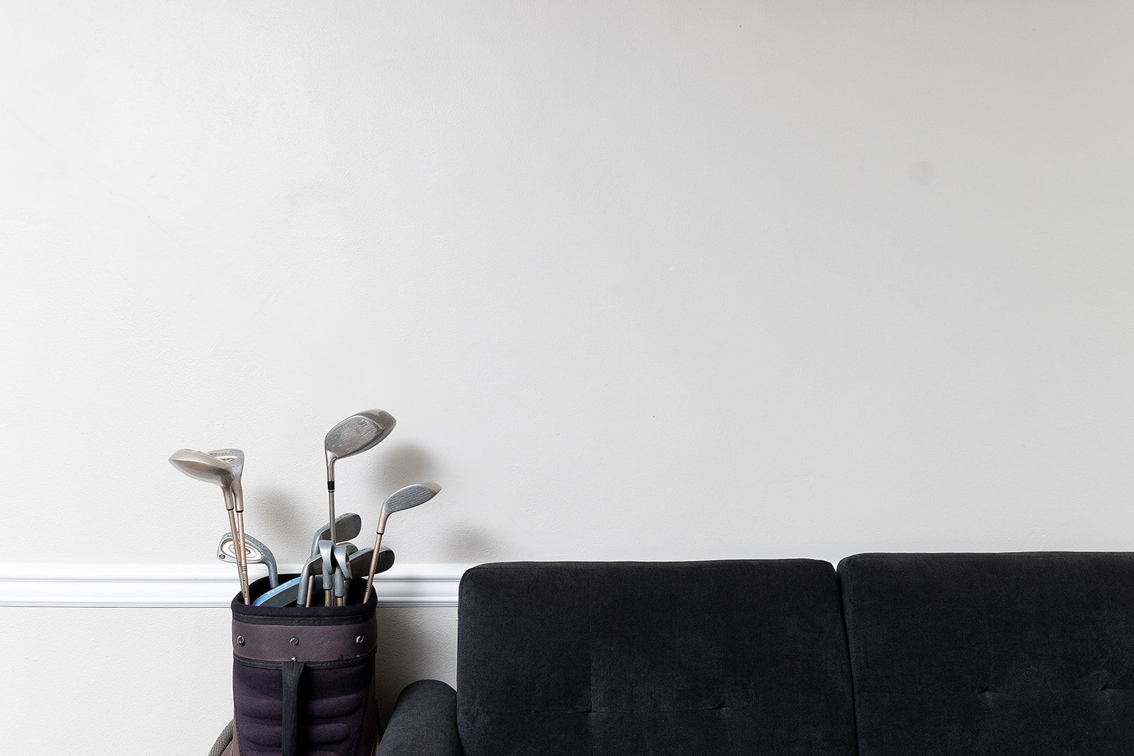 A black sofa against a plain wall with a bag of golf clubs on the floor.