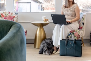 A person sitting on a floral-patterned chair with a laptop, a small table with a cup, and a dog on the floor.