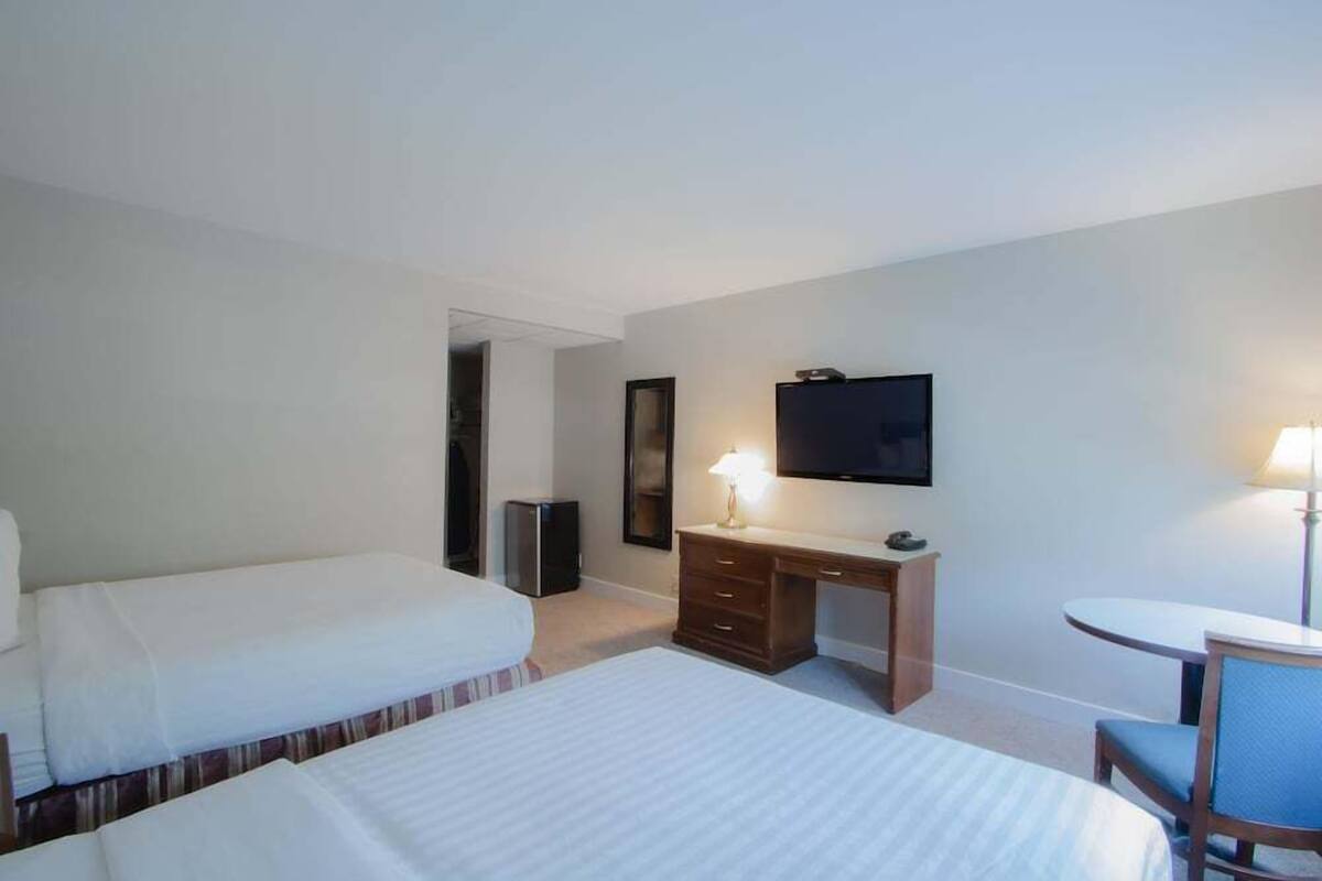 A hotel room with two beds, a desk, a chair, a TV, and a lamp.