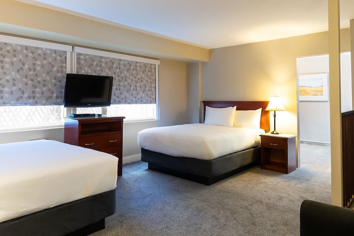 A hotel room with two beds, a TV, a bedside table with a lamp, and a window with patterned curtains.