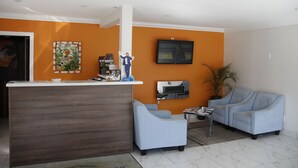 Un área de recepción con un escritorio de madera, dos sillones azul claro, una mesita con revistas, una planta en maceta y un televisor de pantalla plana montado en la pared.