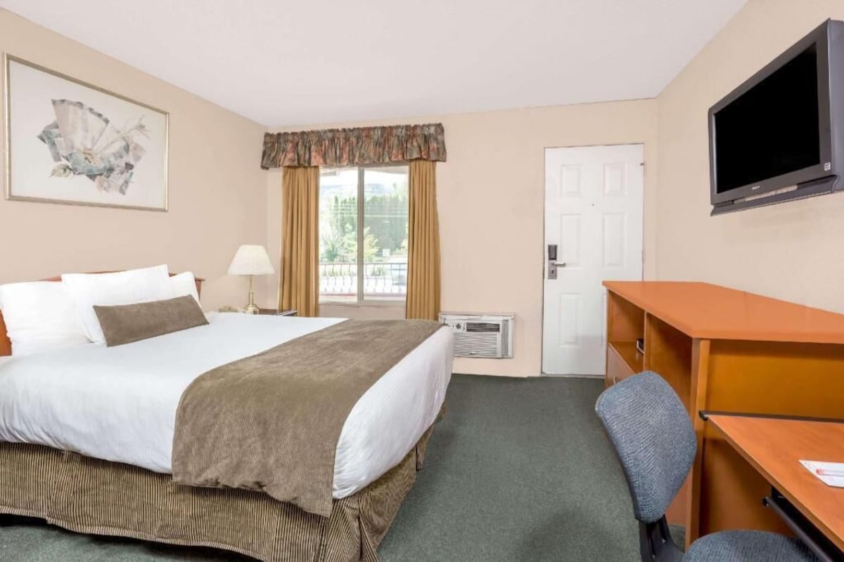 Habitación de hotel con cama, escritorio, silla, televisor, aire acondicionado y ventana con cortinas.