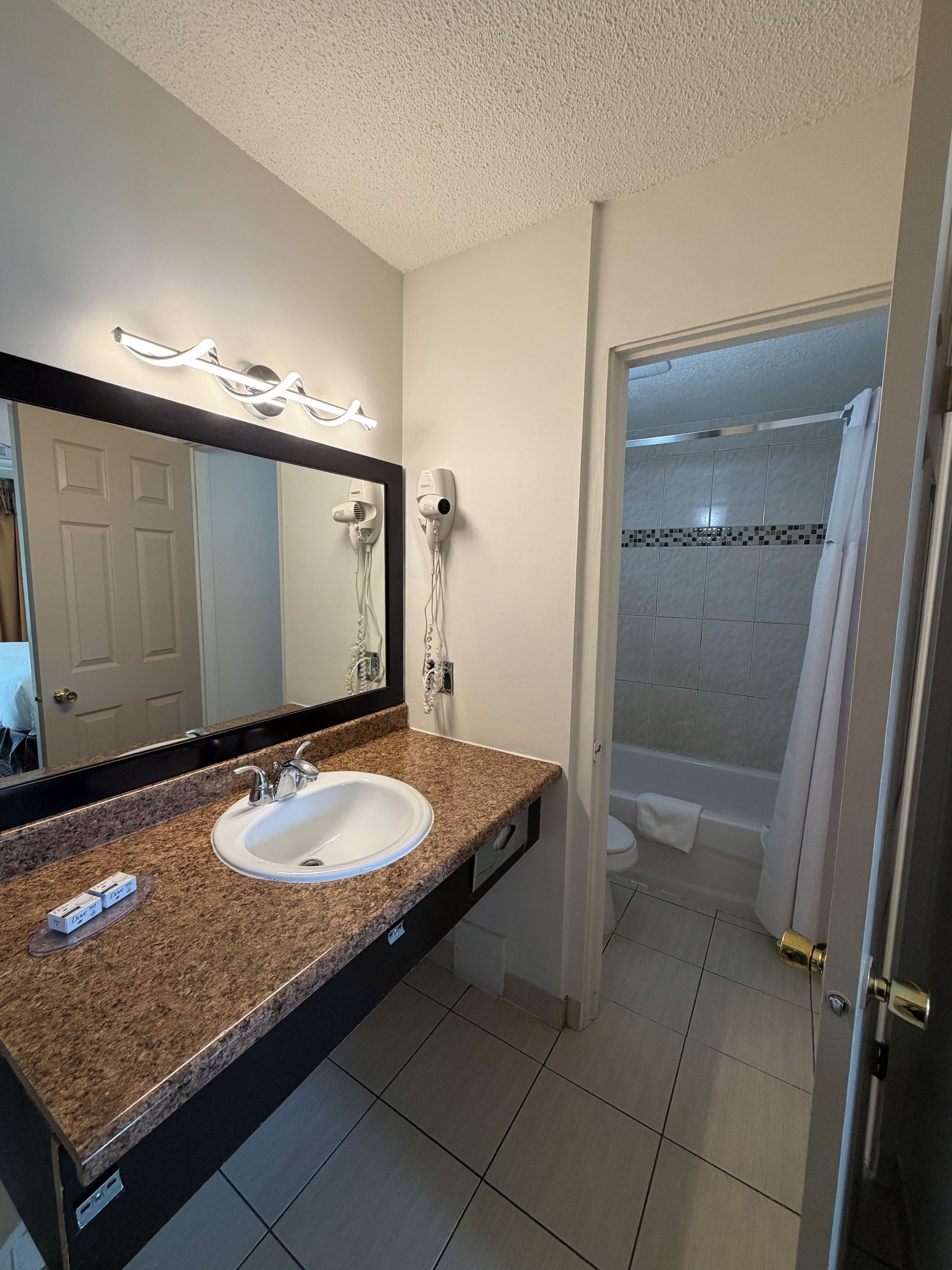 Une salle de bain avec un lavabo doté d’un plan de travail en granit, un miroir avec éclairage intégré, un sèche-cheveux et un espace douche.