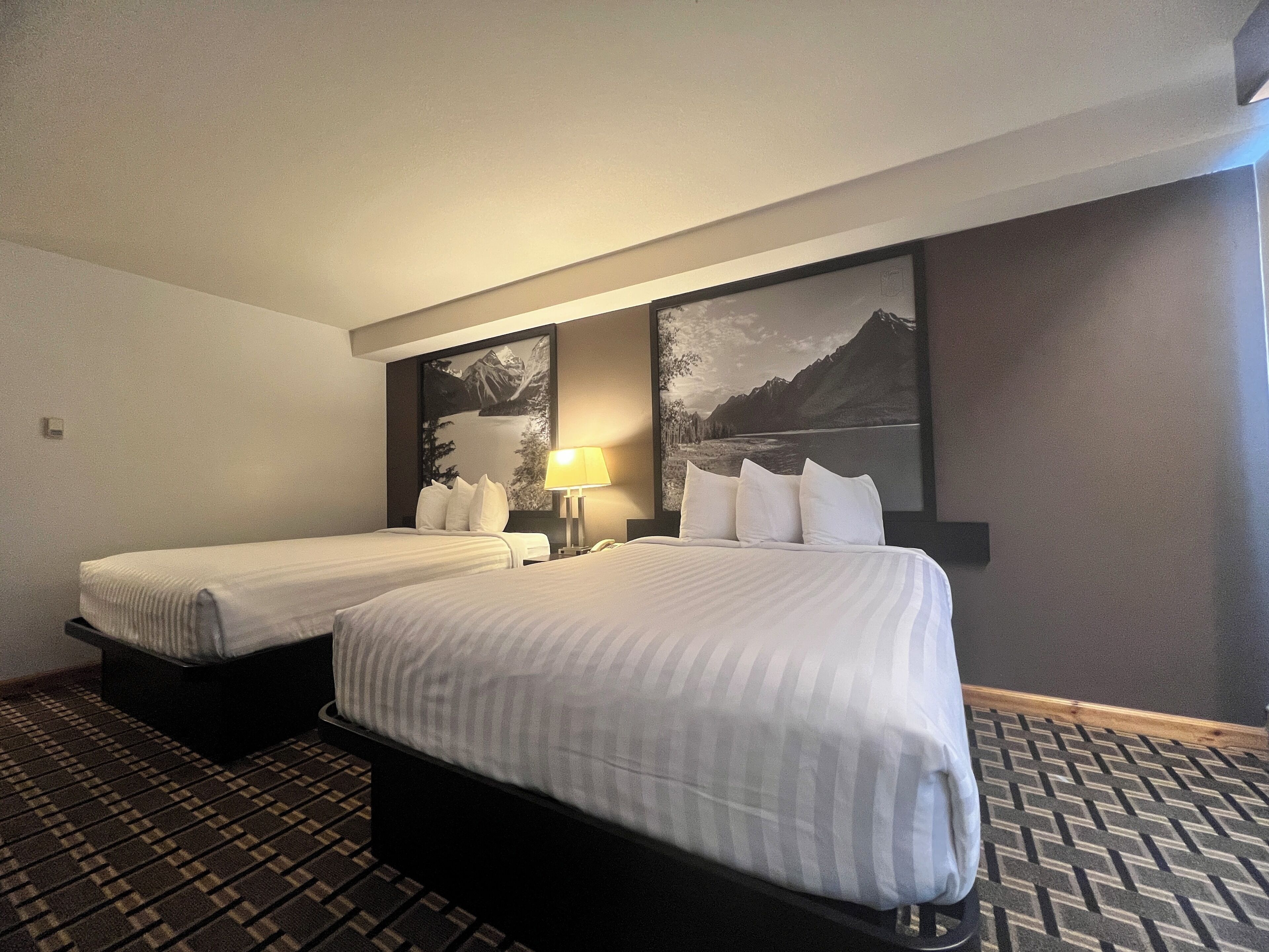 A hotel room with two beds, a large framed picture of a mountain landscape, a bedside lamp, and a patterned carpet.