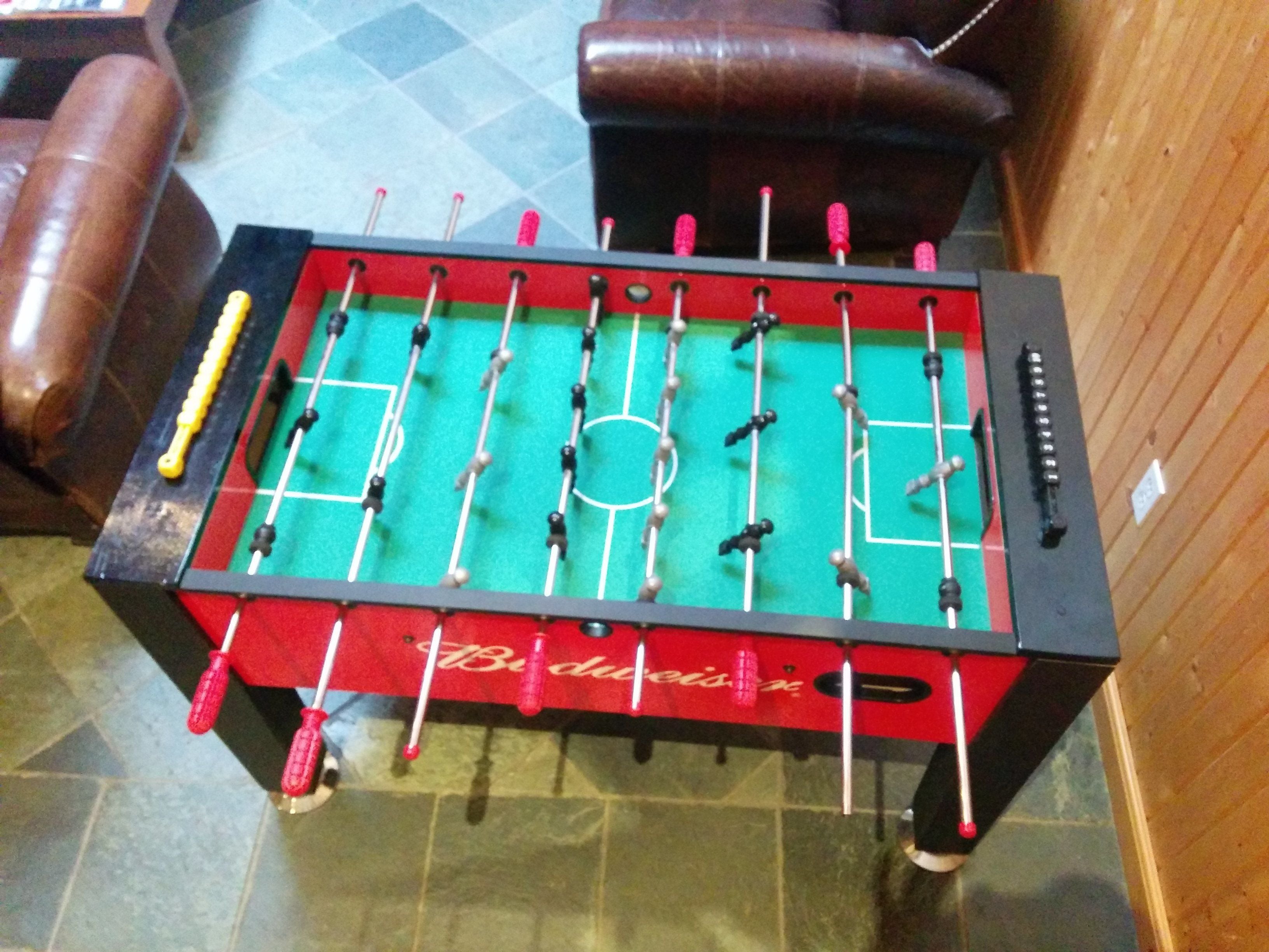 Uma mesa de futebol de mesa com superfície de jogo vermelha e verde, traves pretas e vermelhas, e uma toalha de mesa vermelha com o logotipo Budweiser.