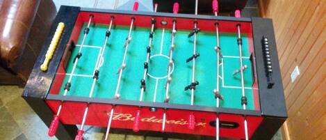 Uma mesa de futebol de mesa com superfície de jogo vermelha e verde, traves pretas e vermelhas, e uma toalha de mesa vermelha com o logotipo Budweiser.