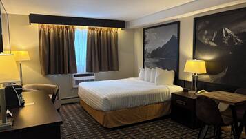 A hotel room with a bed, a desk, a chair, a lamp, a window with curtains, an air conditioner, and a large framed picture on the wall.